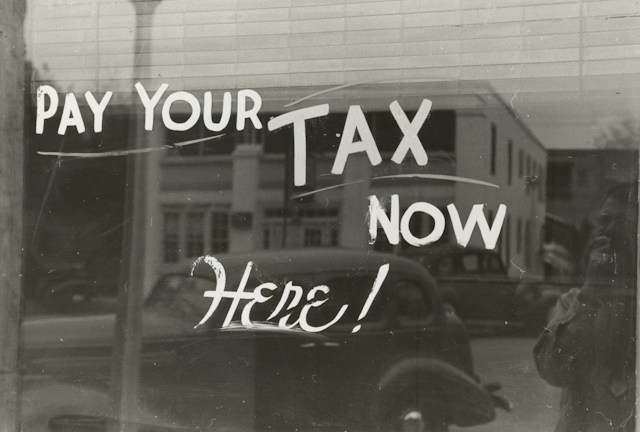 Black and white photo of a window with Pay your tax now! handpainted on it.