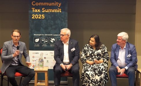 A panel of speakers including three men and a woman. A banner behind reads Community Tax Summit 2025. The man on the left is holding the microphone and everyone is looking at him.